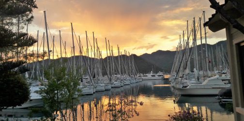 Flotilla of boats docked in a marina setting with a dramatic golden sunset over the mountains beyond reflected in the water