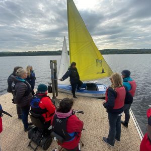 Participants gather around a Sailability yacht with a yellow sail to receive an induction before heading out on the water