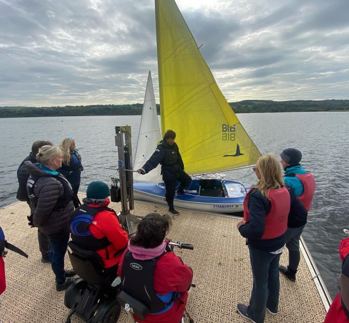 Participants gather around a Sailability yacht with a yellow sail to receive an induction before heading out on the water
