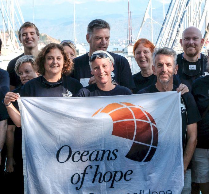 Image of the Oceans of Hope team all wearing dark blue tee shirts and holding up a large white Oceans of Hope flag with masts of yachts in the background