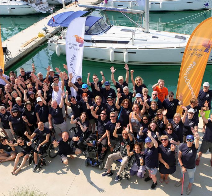Ariel view of a big group of challenge participants standing and some in wheelchairs all waving and smiling to camera with orange and white flags and a yacht in the background