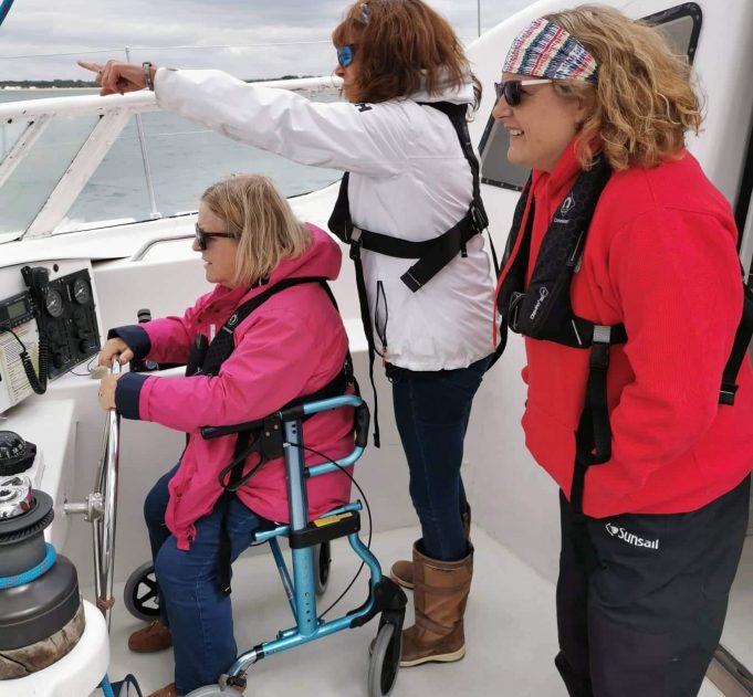 Nicola on board Spirit of Scott Barder catamaran wearing a red sailing jacket and talking to a sailing participant in a wheelchair who is at the helm and another sailor pointing the direction
