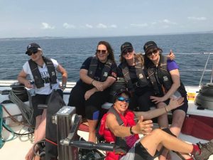 A group of participants pose for the camera in a cockpit of a yacht with calm seas and blue skies behind them