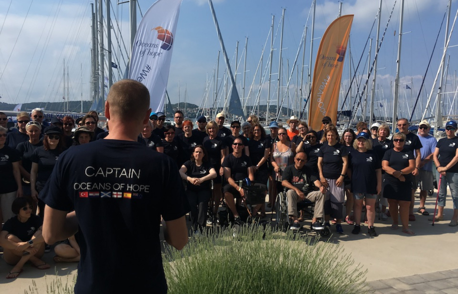 A big group of participants some in wheelchairs are looking at Mikkel taking who is taking a group photo of them all in their blue oceans of hope tee shirts with orange and white flags and the masts of yachts in the background