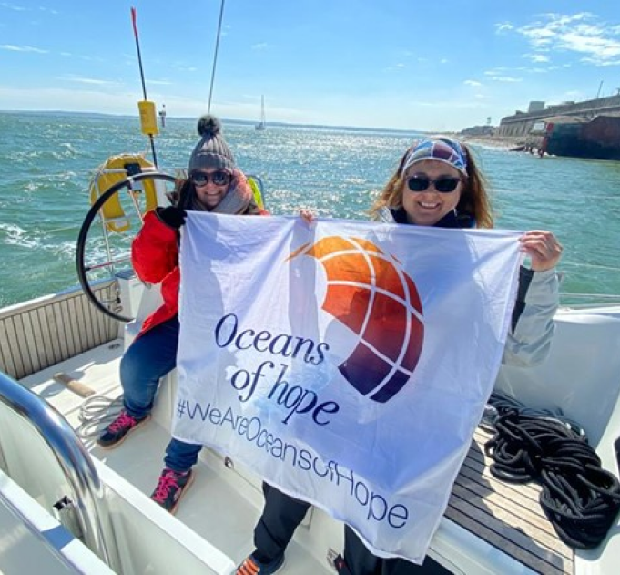 Nicola and a fellow sailor holding up an Oceans of Hope flag on the deck of a yacht which is sailing on a light blue sea with the sky behind