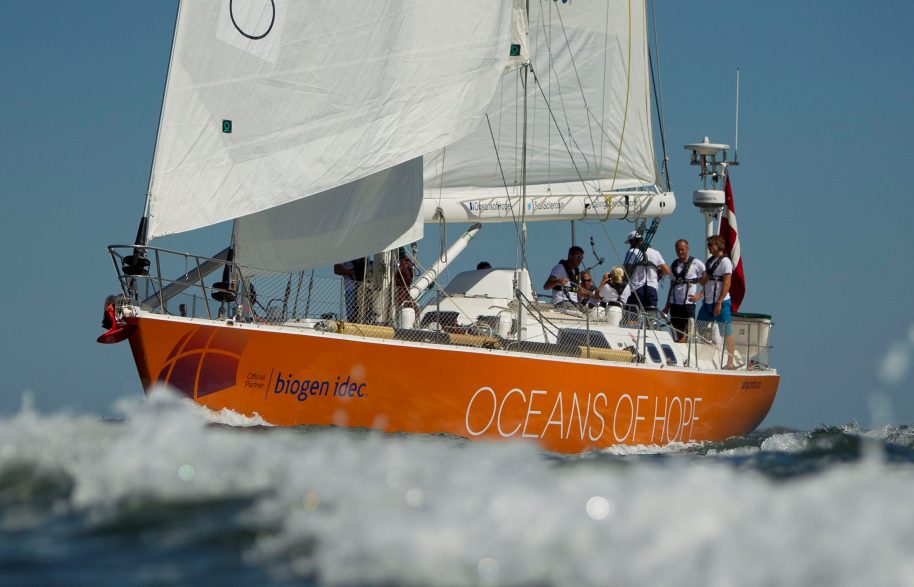 Orange hull of the Oceans of Hope yacht racing across the waves against a blue sky with white surf in the foreground