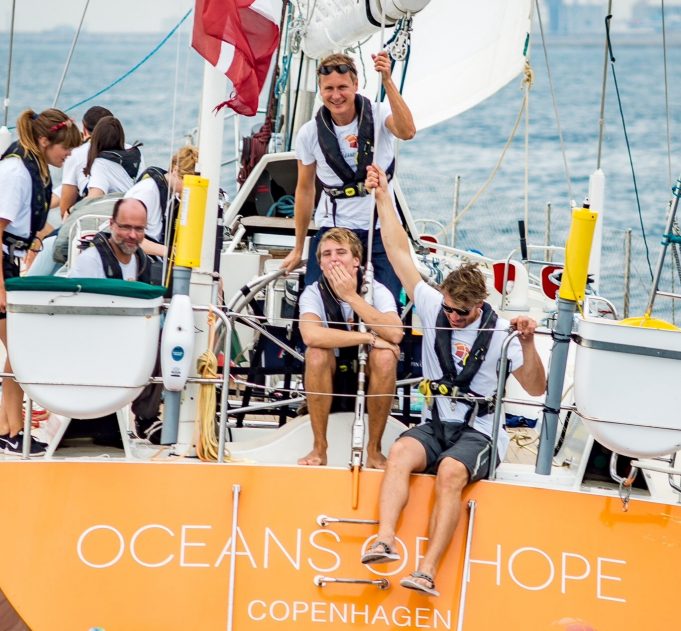 Sailors on board Oceans of Hope yacht with orange coloured stern carrying the boats name and ocean in the background