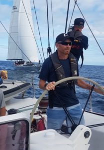 A participant takes the helm of a yacht with another yacht visible in the background under sail