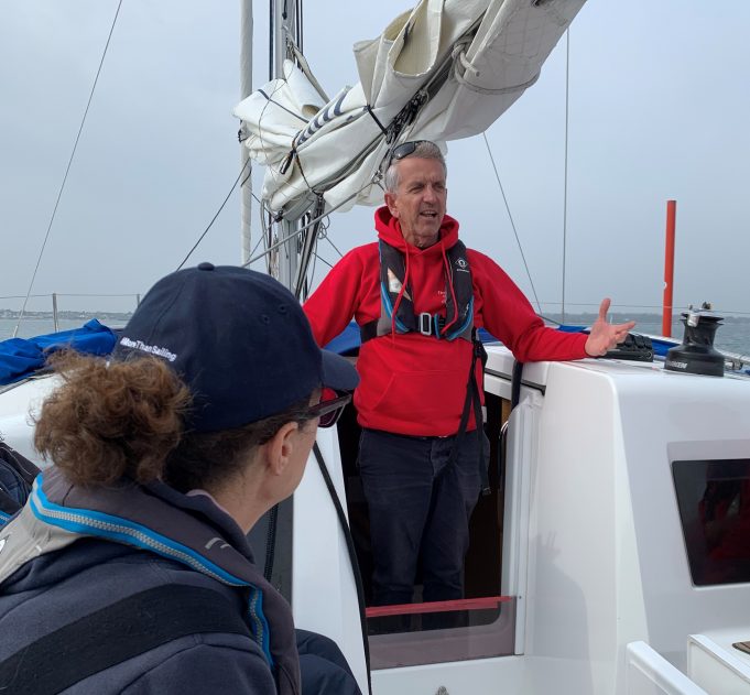 Robert standing on the deck of a yacht in a red sailing jacket talking to the crew